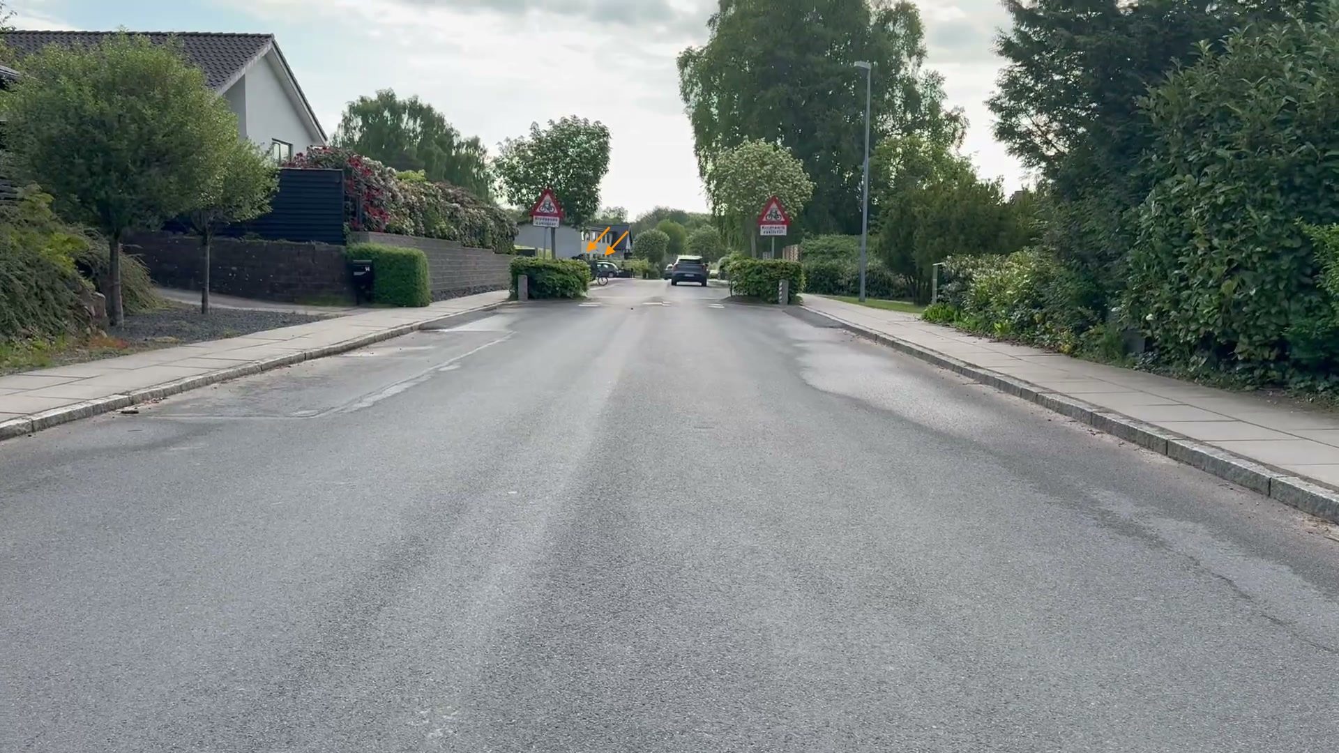 Bilist-perspektiv på Vesterlundvej. To skolebørn på cykel ved cykelstiens udmunding er kun marginalt synlige som mørke konturer over chikane-bedets løv