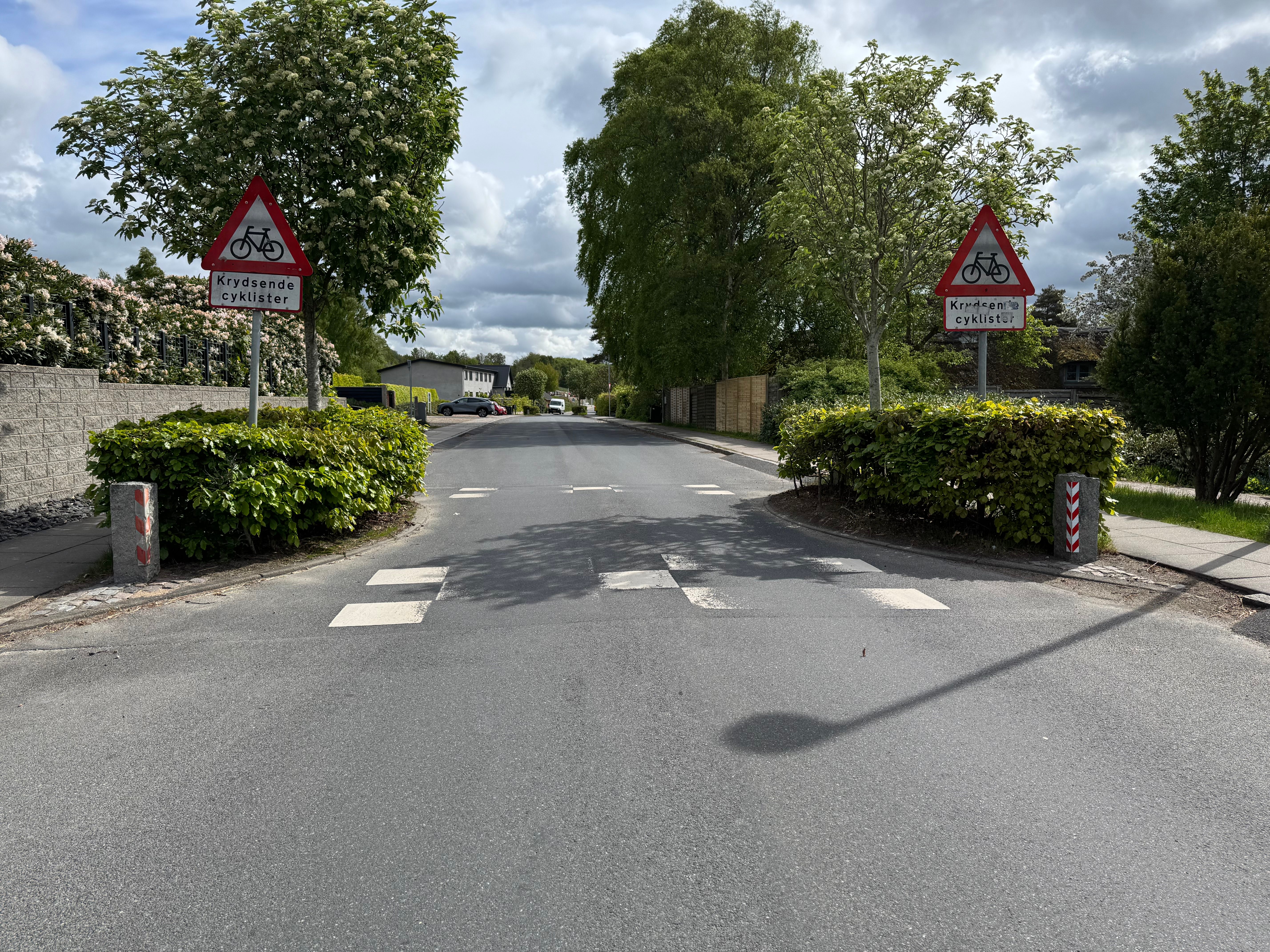Sigtelinje fra cirka 30 m vest for krydset. Skiltene og helleanlægget er nu tydelige, men cykelstiens åbning er stadig skjult bag hækbeplantningen