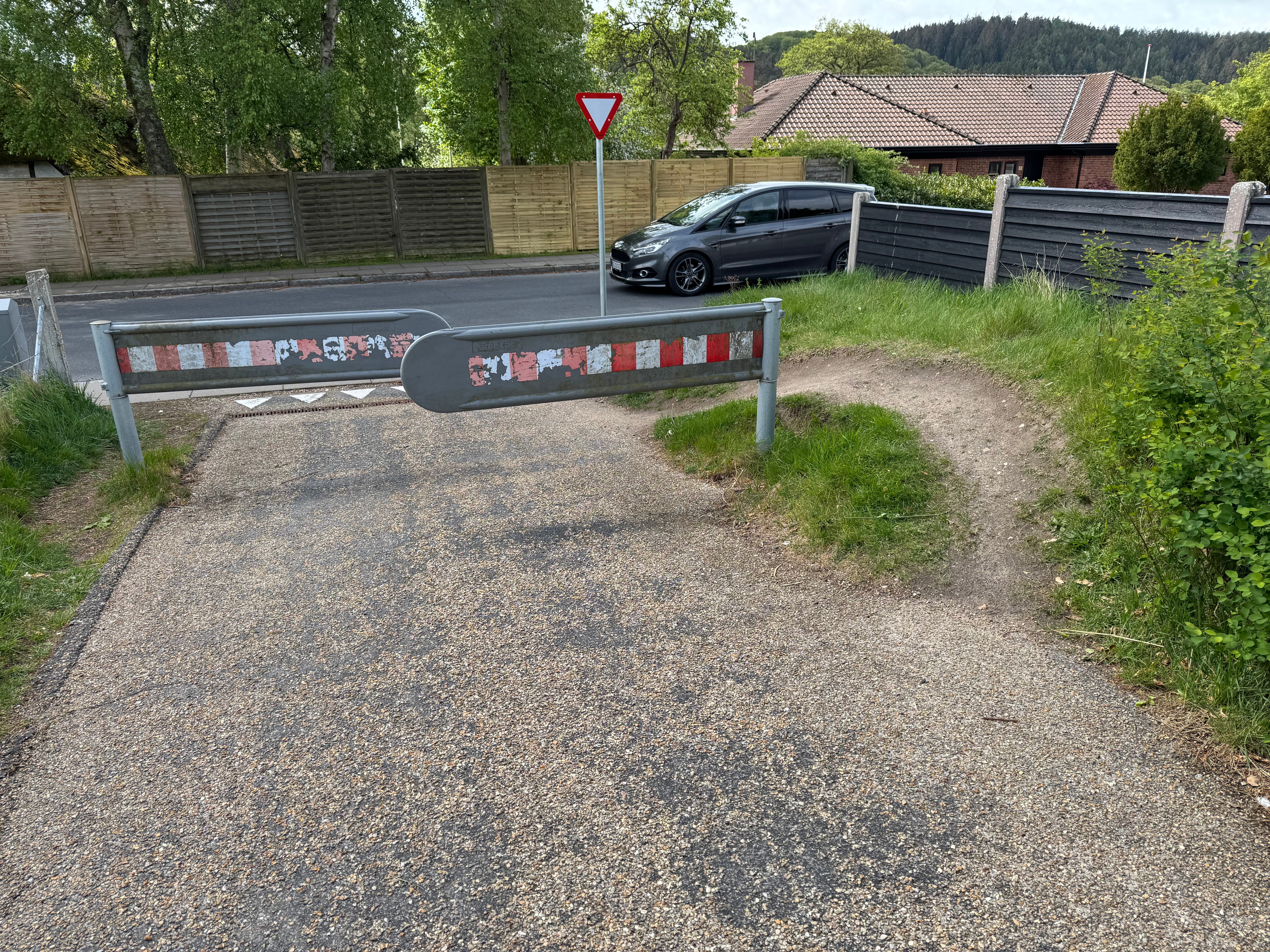 Cyklistens perspektiv mod Bom 1, den sydligste bom. To selvlukkende spærrebomme stillet forskudt så stien spærres i zigzag. Til højre løber et tydeligt slidt spor i jorden uden om hele installationen. I baggrunden Vesterlundvej og et 'vigepligt'-skilt
