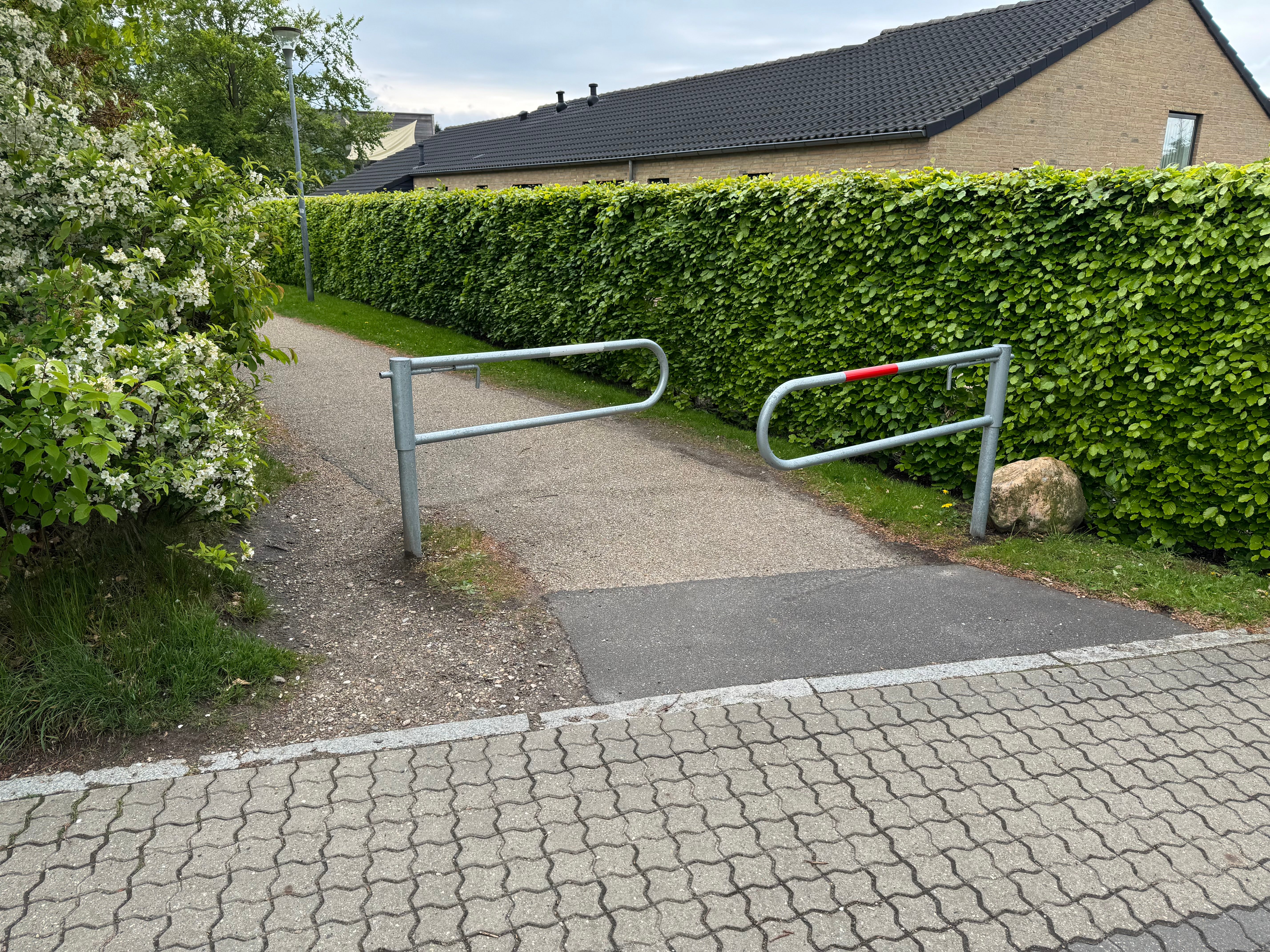 Bom 3, den drejelige spærrebom i galvaniseret stål, ses i forskydning. Langs højre side af stien løber en jord-zone uden græs umiddelbart op ad asfaltkanten